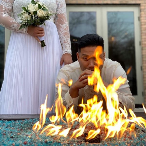 A man in a white shirt looks at a fire pit, with a woman in a white dress behind him.