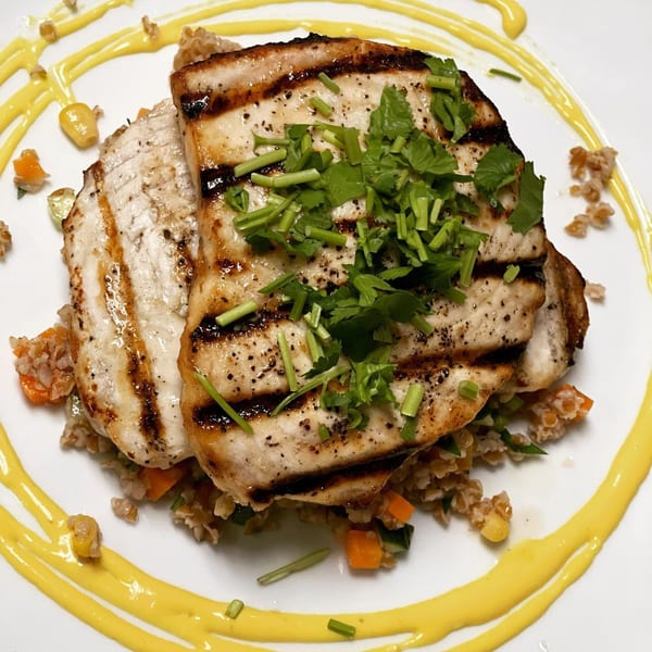 Grilled fish fillets with herbs on a bed of grains and vegetables, drizzled with yellow sauce.