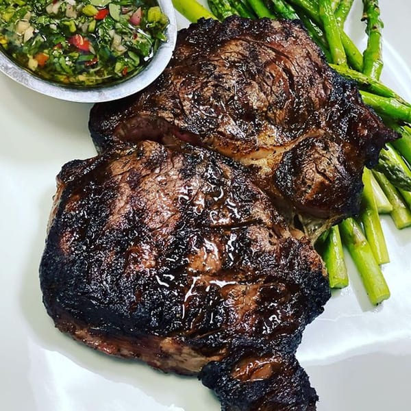 Two grilled steaks with a side of chimichurri sauce and green asparagus on a white plate.