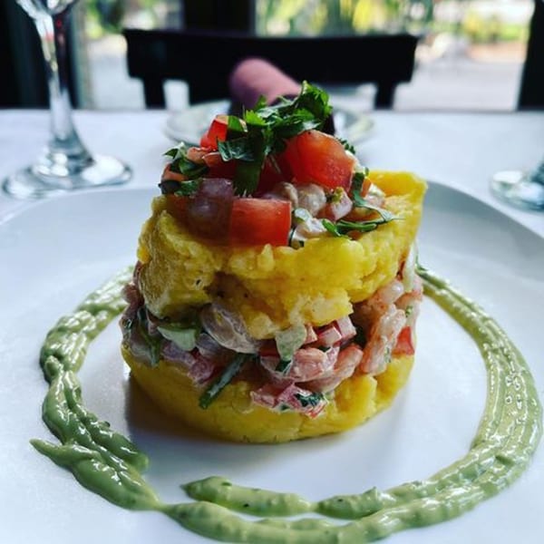 A vibrant Latin-American dish with yellow mashed potato, shrimp, tomatoes, and green sauce on a white plate.
